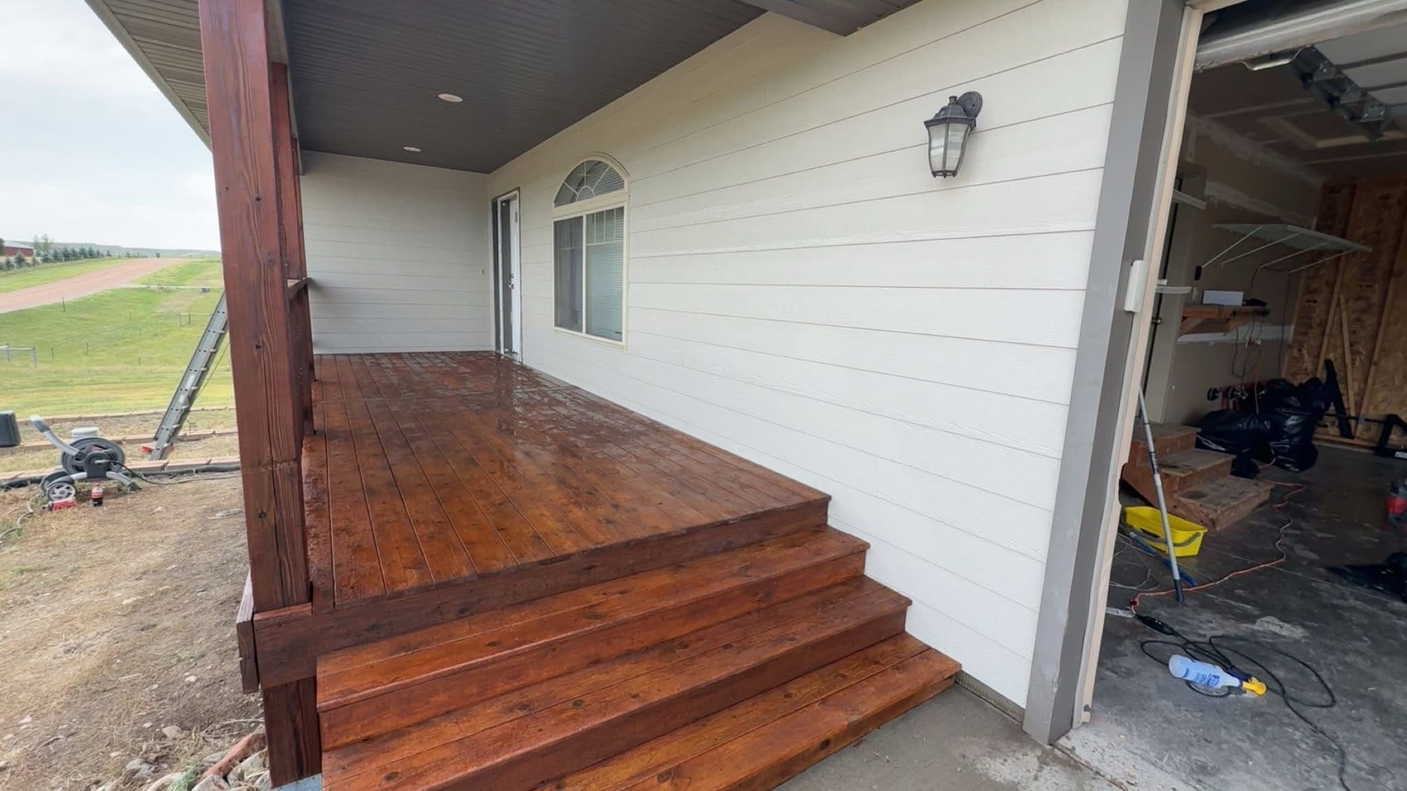 A covered porch with the deck and its steps freshly stained, against white painted siding, with a wall lantern and the garage bay open alongside