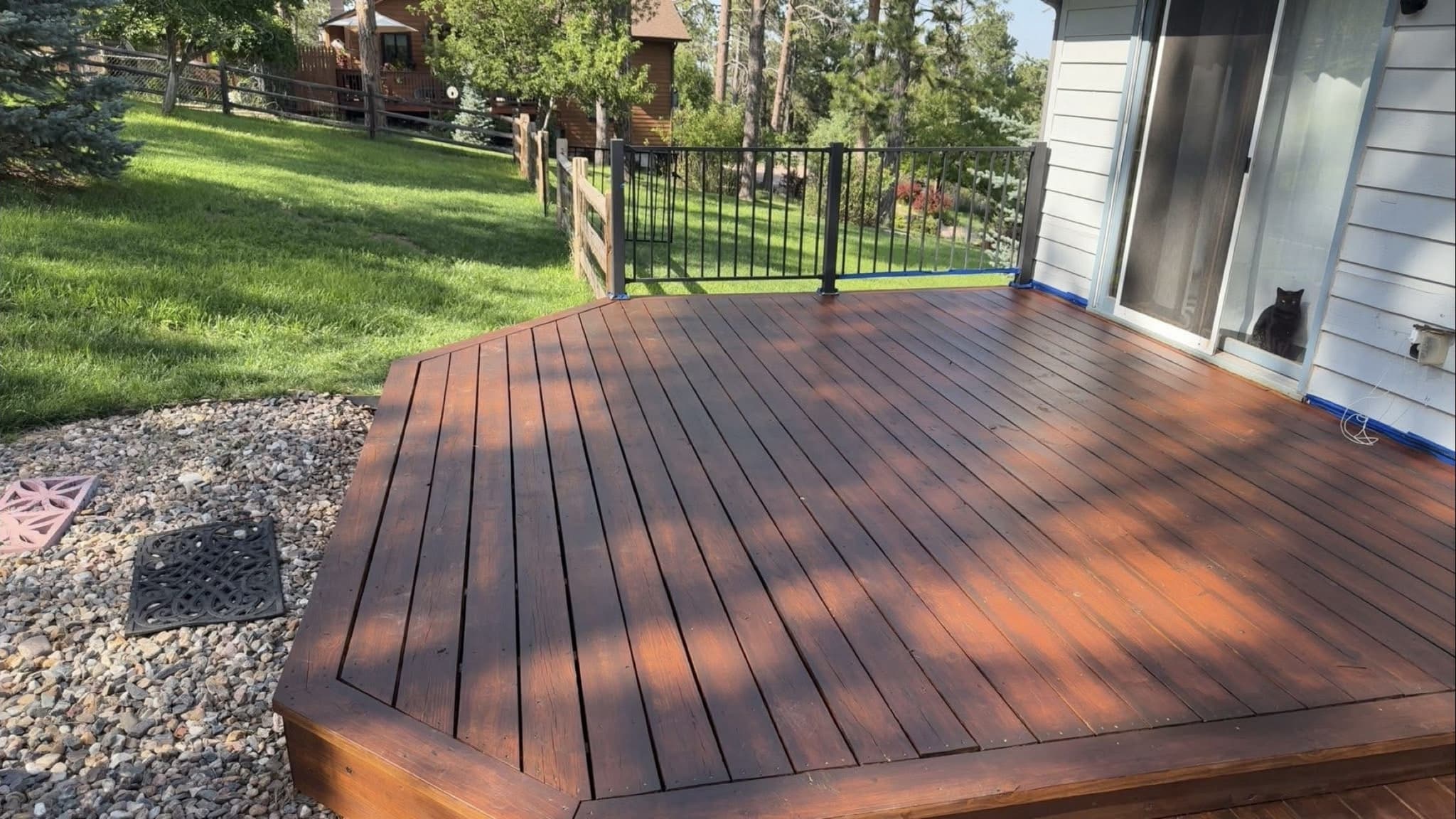 A deck stained a dark red-brown across every board, with a black iron rail at the far edge and lawn and pines beyond