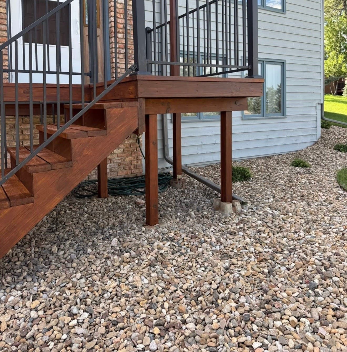 Stained deck stairs, posts and framing with a black iron rail, meeting brick and lap siding above a gravel bed