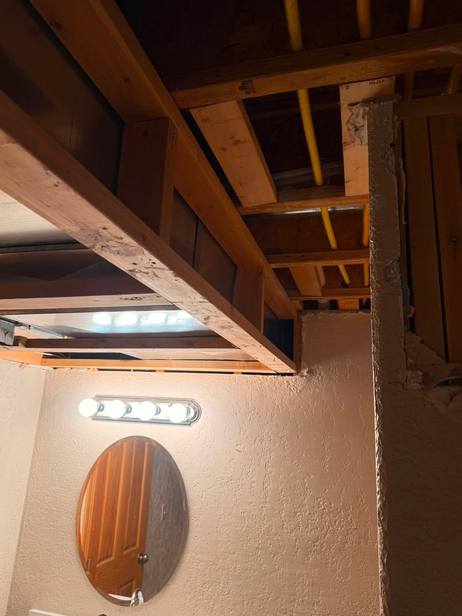 A bathroom ceiling opened to the joists directly above a vanity light and oval mirror, framing and pipework exposed