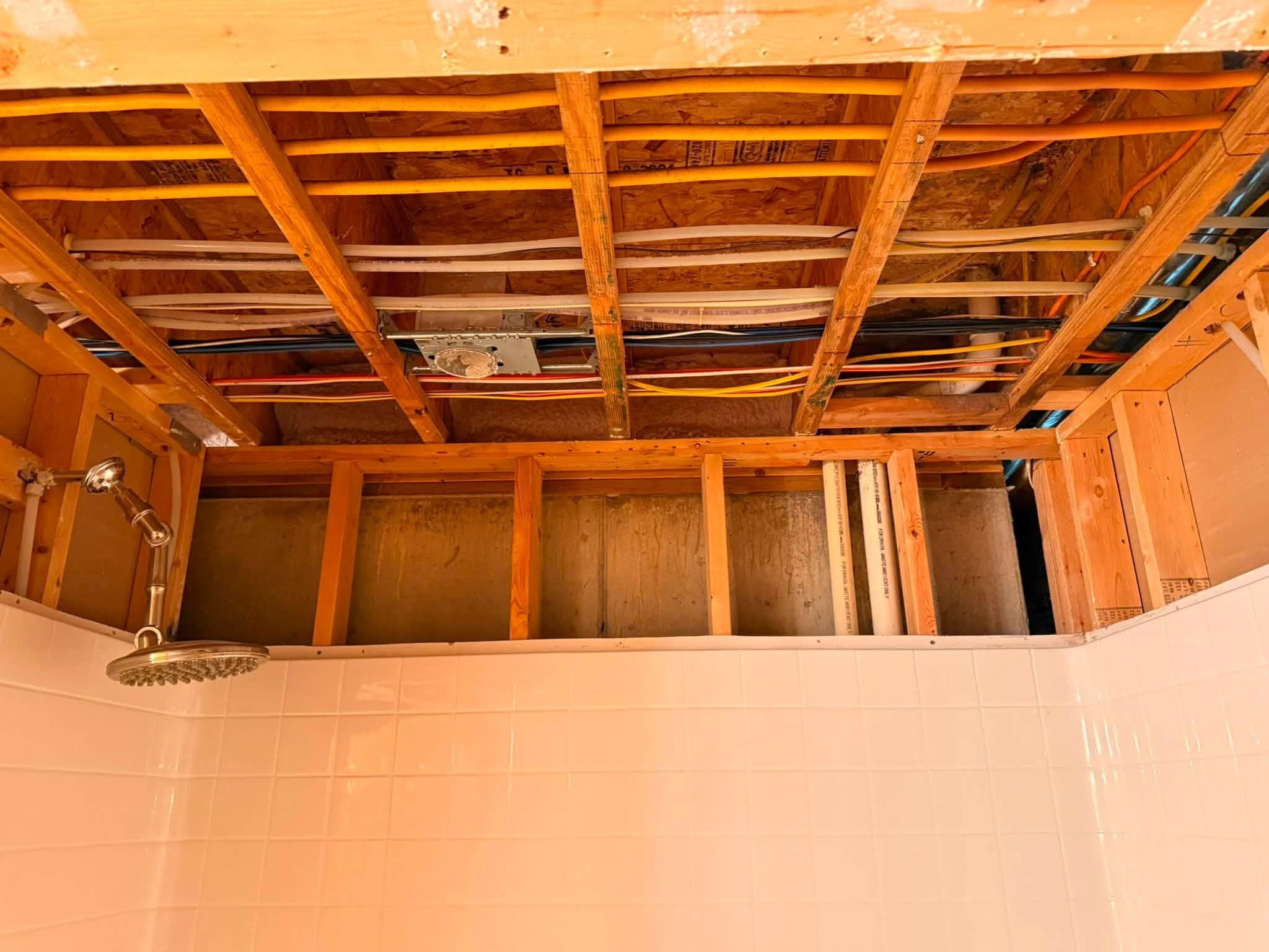 A ceiling stripped back to bare joists above a tiled shower, with the wiring, pipe runs and shower arm left in place