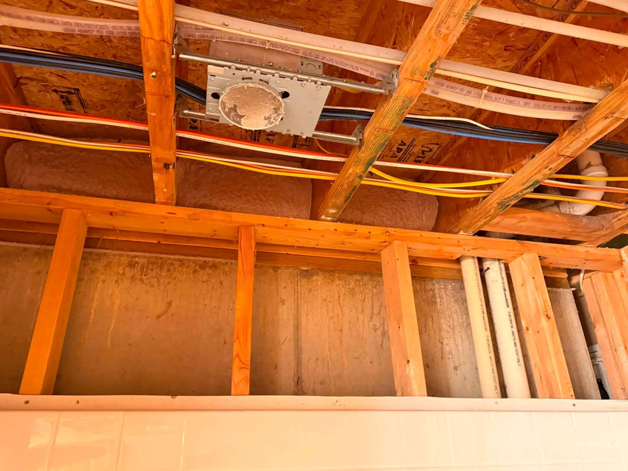 Open ceiling joists carrying wiring, flexible duct and an electrical box, above a tiled wall