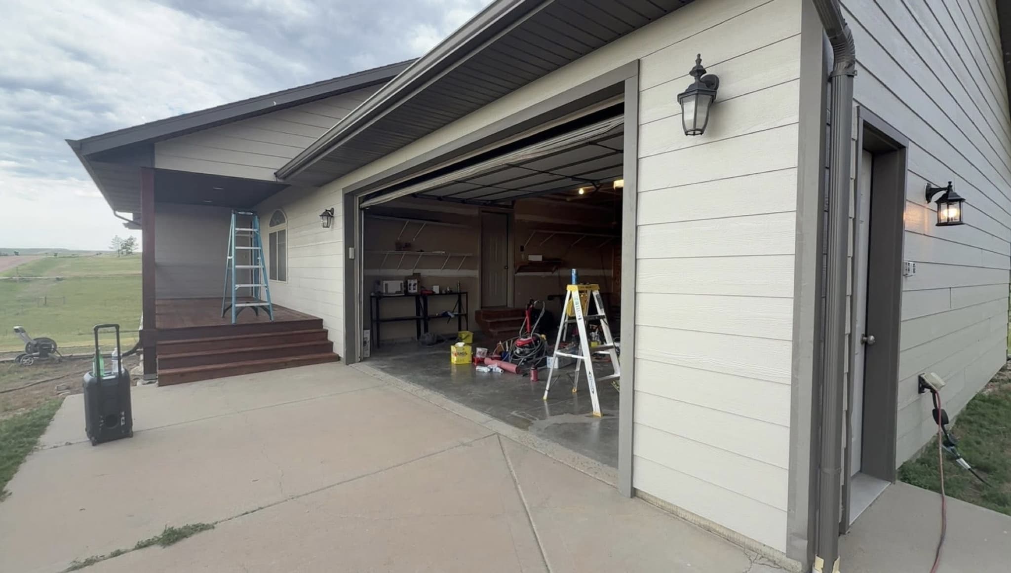 A single-story house with pale lap siding, its garage door open on a step ladder and painting equipment, a second ladder standing on the freshly stained dark deck alongside, under an overcast sky.