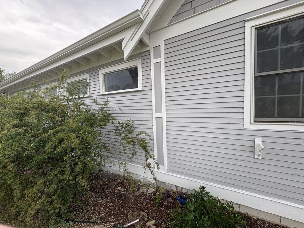 Gray painted lap siding with white trim, a gable bracket and a picture window, shrubs along the foundation