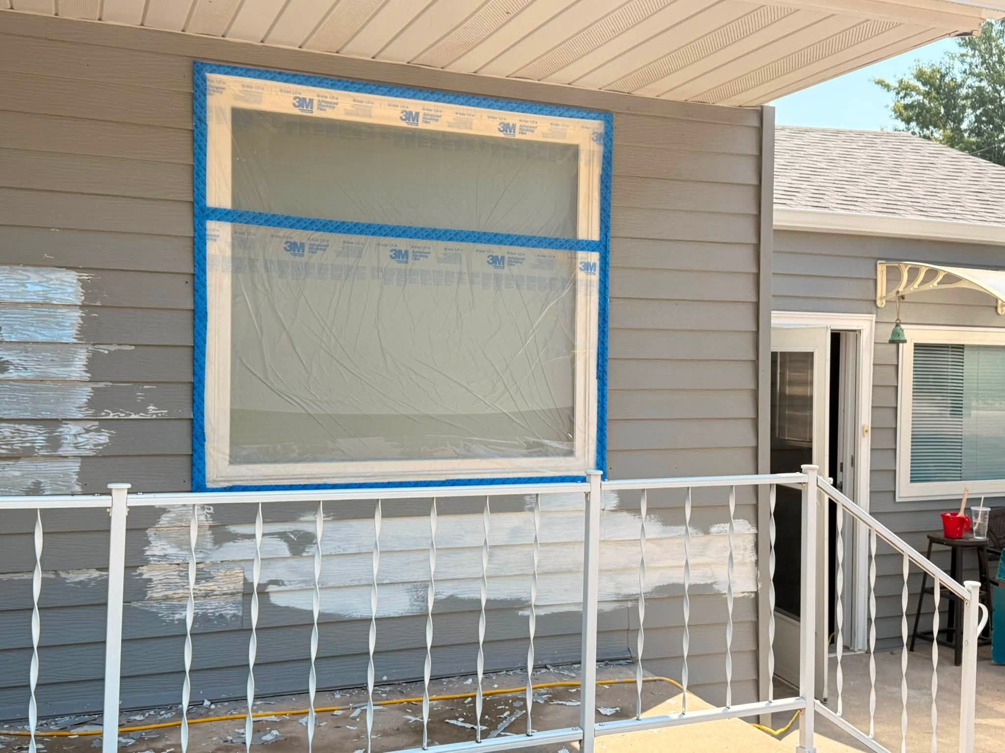 Preparation stage: a window fully masked with tape and film, siding scraped back and spot-primed, porch railing covered