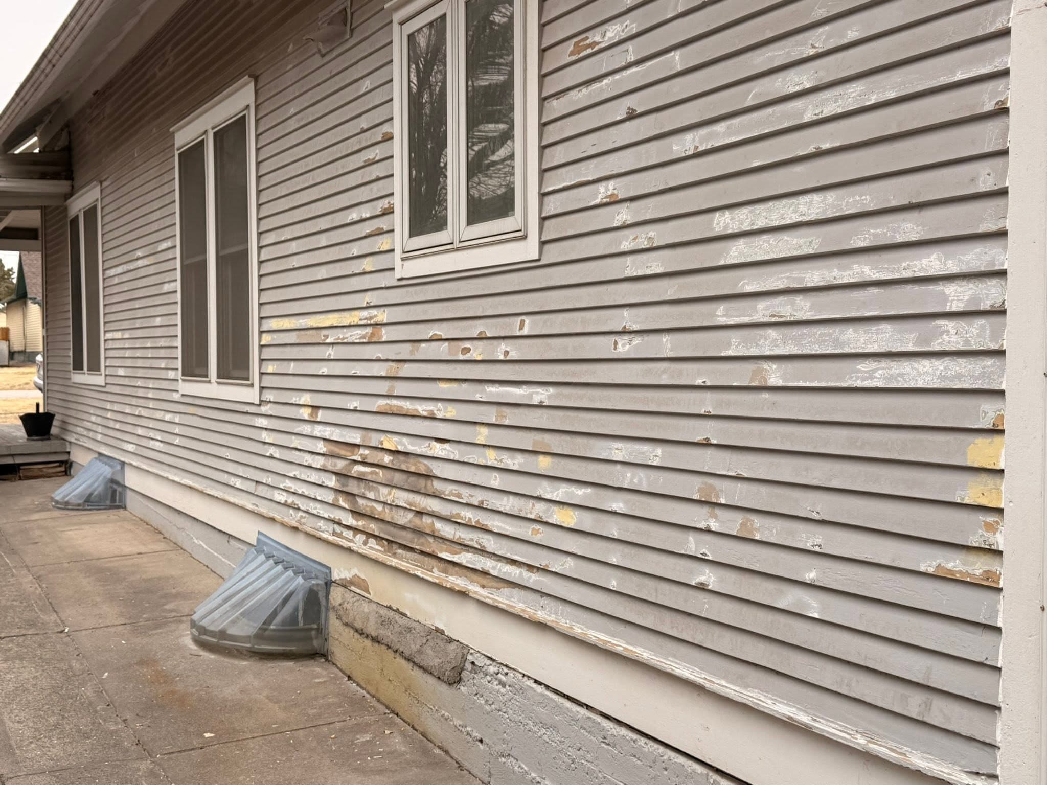 Lap siding part-scraped, the old finish still on the upper courses and bare patches below, with a splash block at the base