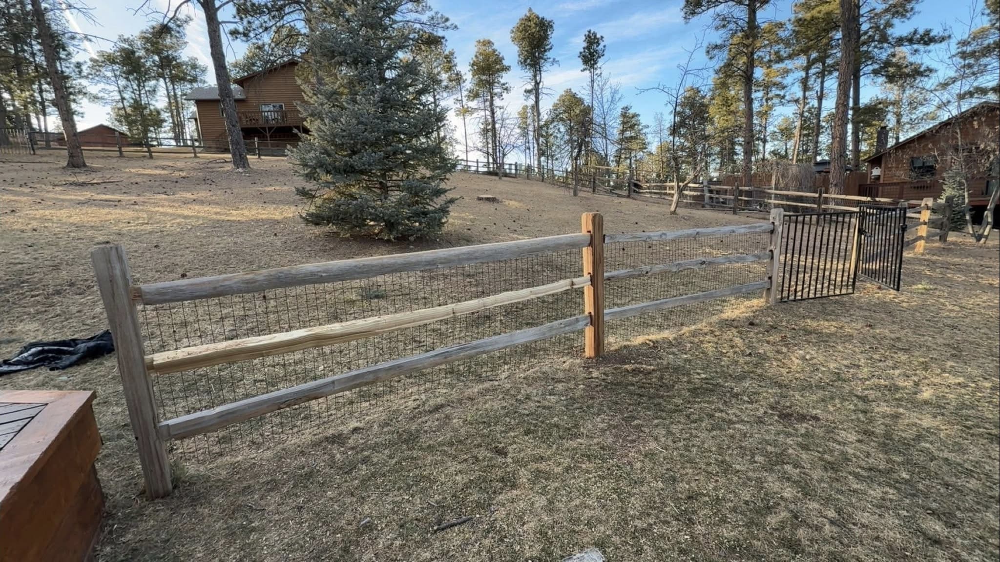 A split-rail fence backed with wire mesh, one rail and post replaced in fresh timber, running along to a black metal gate