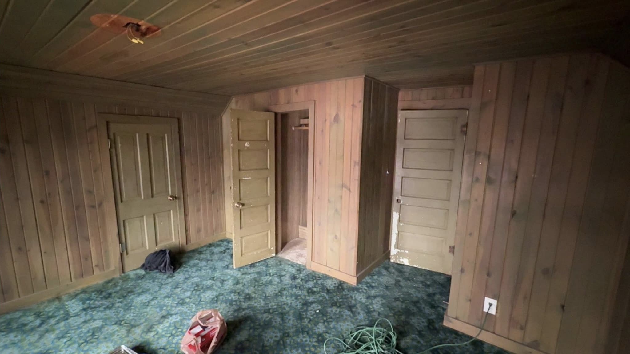 A room before work: knotty timber paneling floor to ceiling, patterned green carpet, and a bare fitting on the plank ceiling