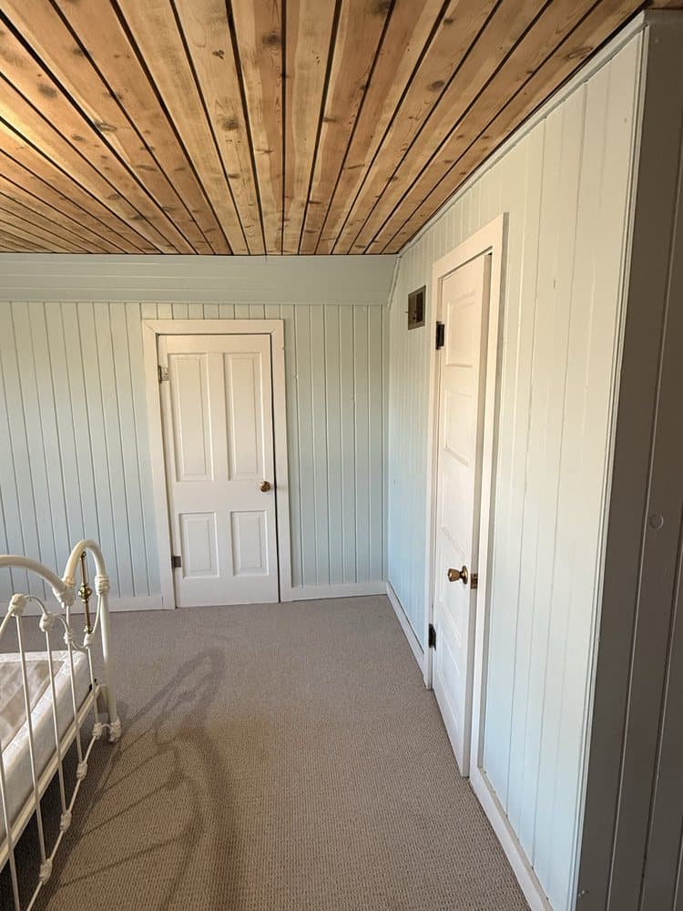 Bedroom with a cedar plank ceiling above pale painted vertical paneling, a white six-panel door and carpet