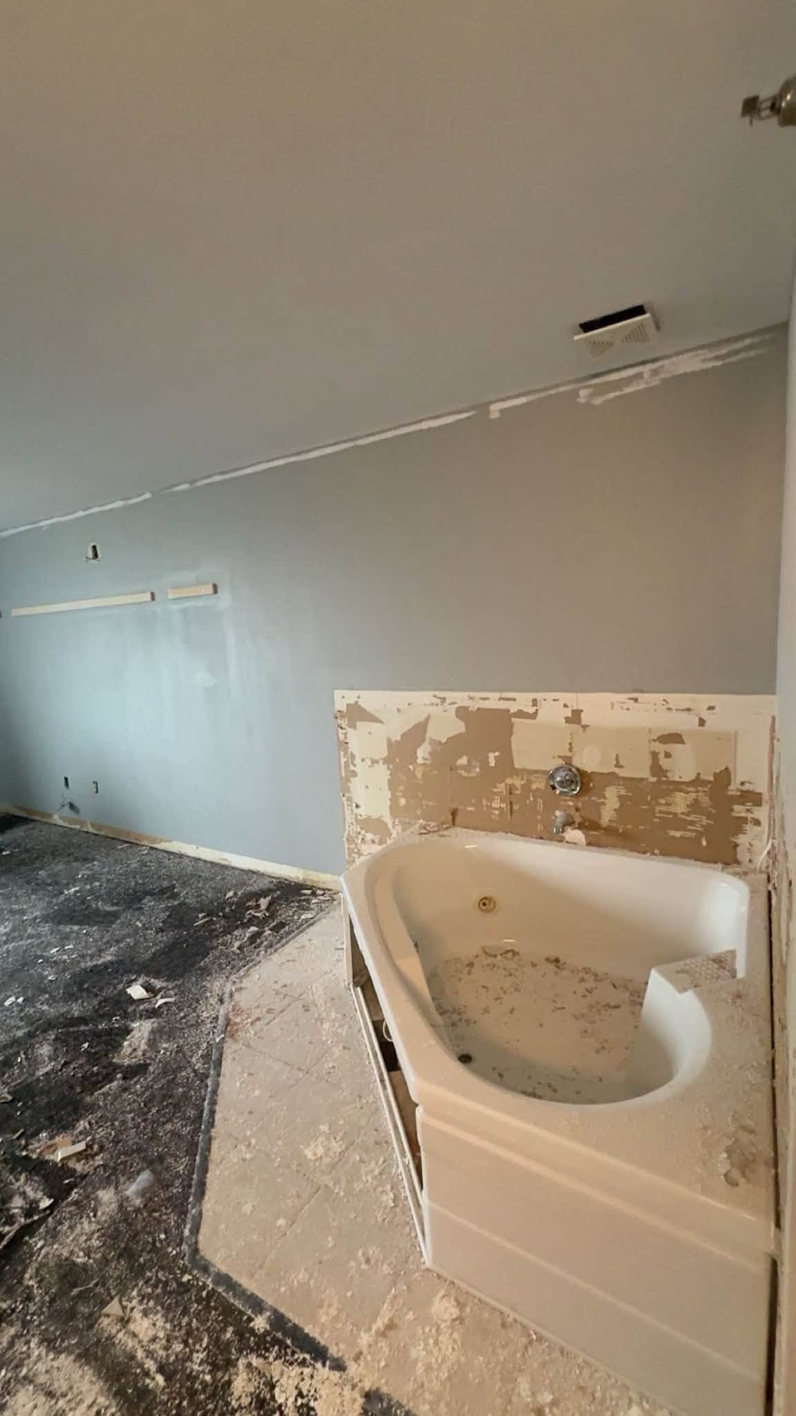 A bathroom stripped out: a corner tub left standing, the flooring taken up to black adhesive and the wall opened alongside