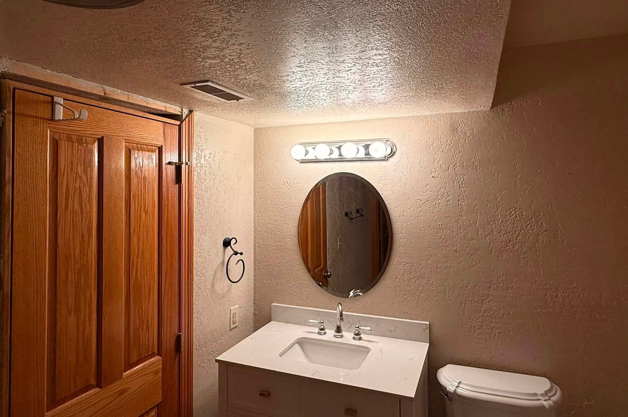 A finished bathroom with an oak door, a basin and vanity under an oval mirror and a four-bulb light bar