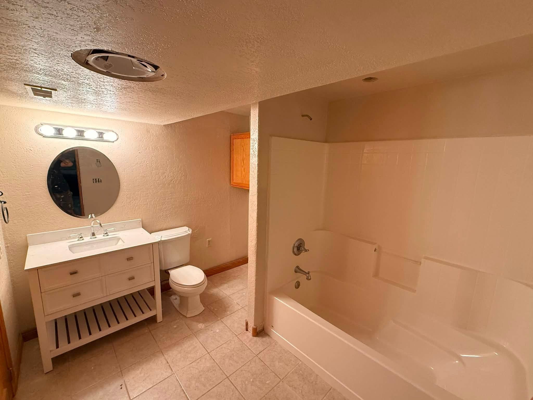 A finished bathroom: a light timber vanity and basin, a WC and a long tub under a tiled surround, on tile flooring