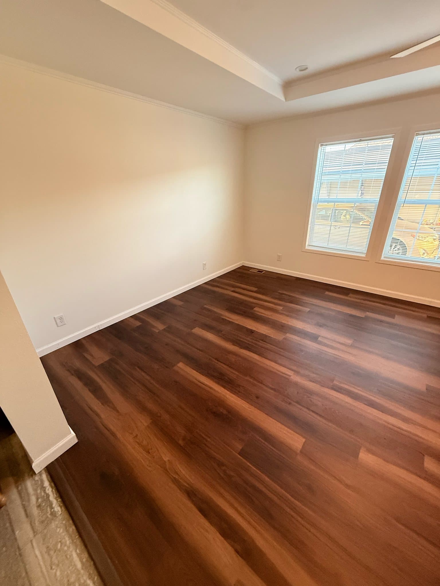 An empty room with cream walls and white baseboards, dark plank flooring running to two windows with blinds in the far corner