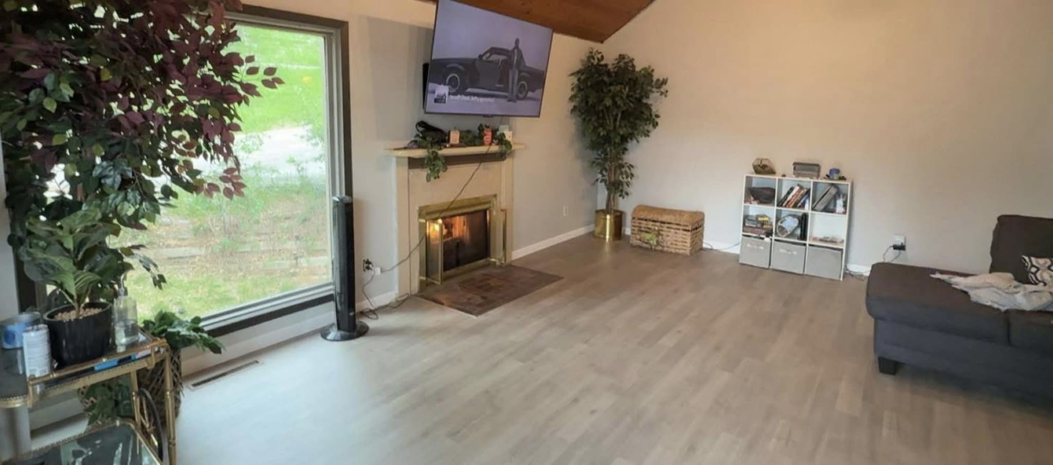 A finished living room with wide plank flooring, a fireplace under a wall-mounted television, and a picture window onto the yard