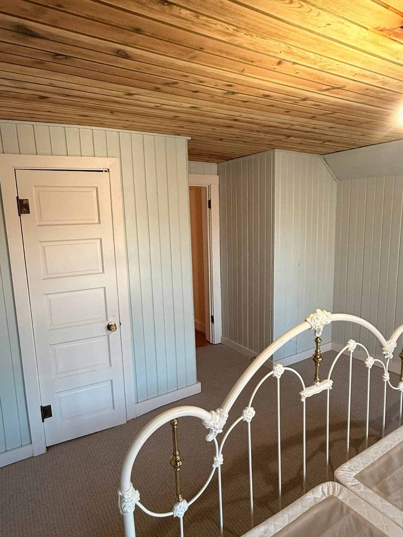 A bedroom with pale painted vertical paneling under a cedar plank ceiling, a white six-panel door and an iron bedstead