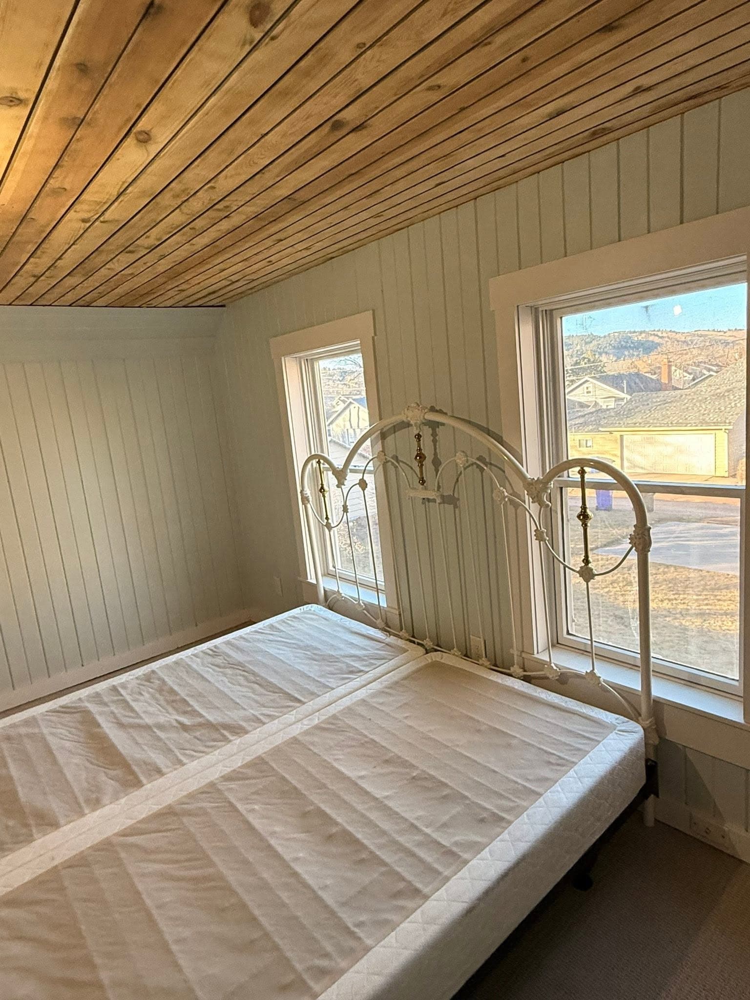 A paneled bedroom under a timber plank ceiling, with a metal bed frame set at the window