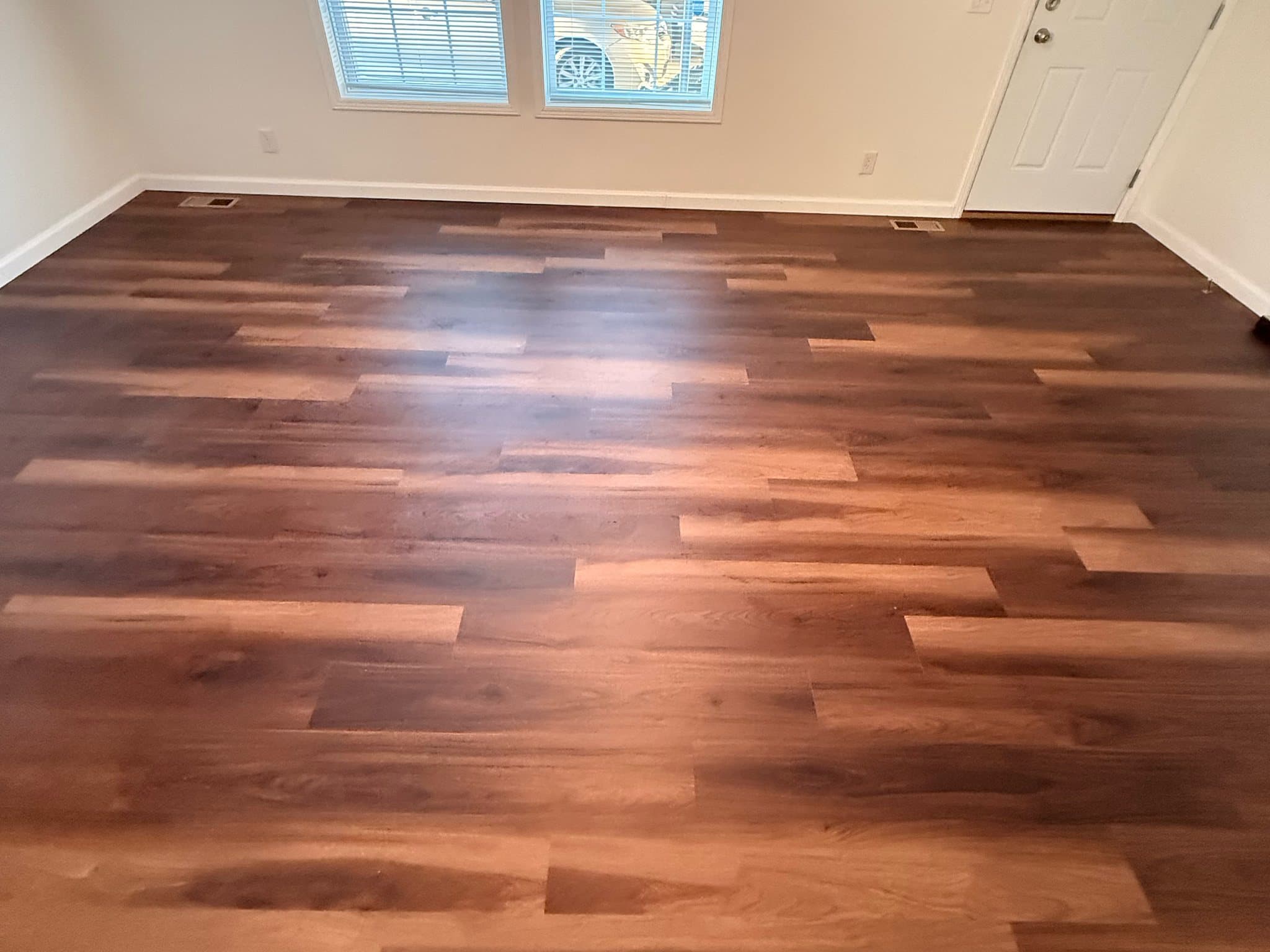 Dark plank flooring across an empty room, running to cream walls, white baseboards, two windows with blinds and a white paneled door