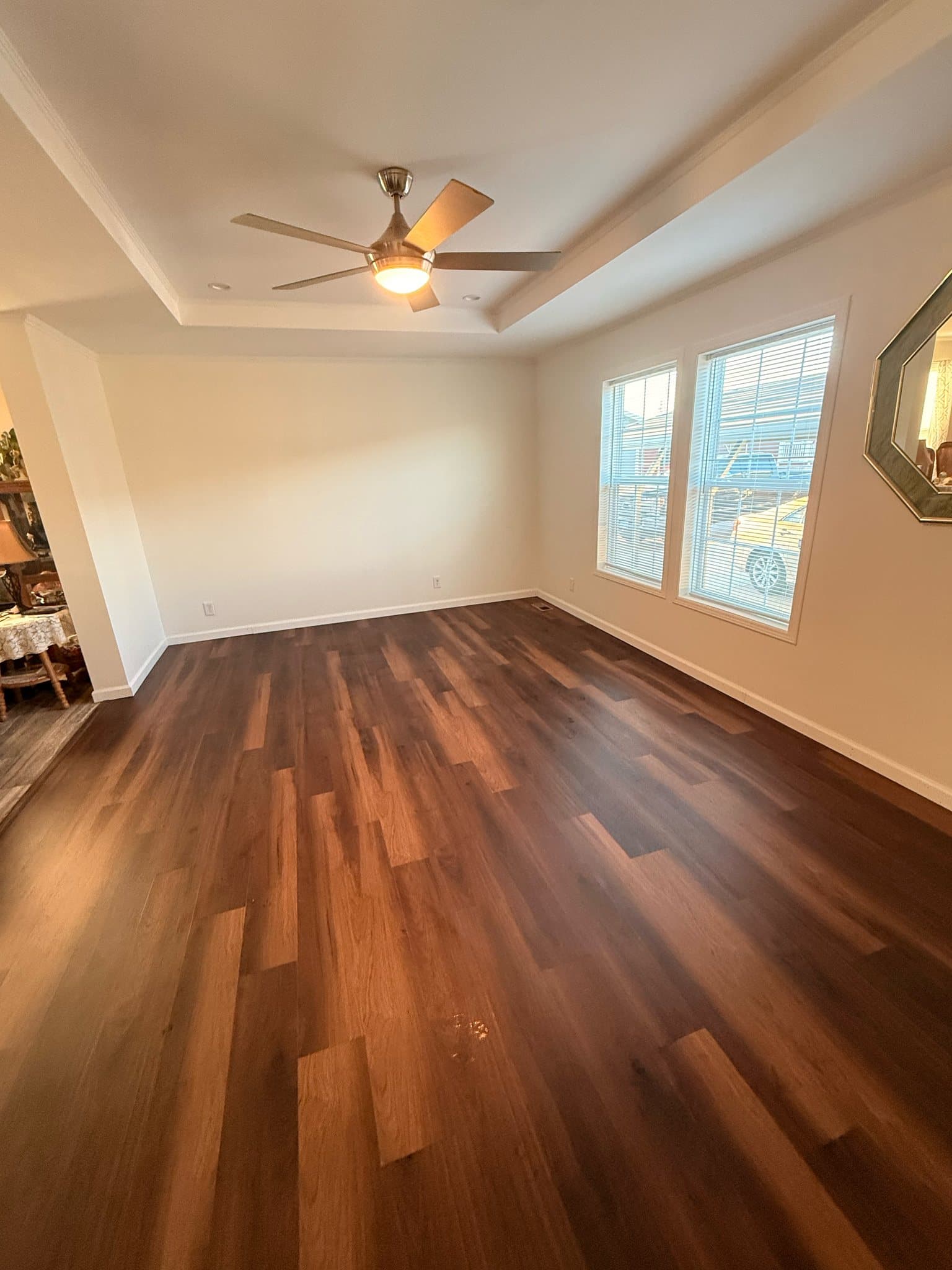 An empty room under a tray ceiling, with cream walls, white baseboards, dark plank flooring and two windows with blinds along one wall